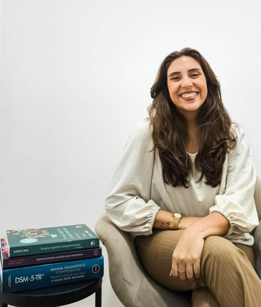 Fotografia profissional da psicóloga clínica Louize Vieira sorrindo. Ao fundo, uma estante de livros, transmitindo segurança, estudo e autoridade em Terapia Cognitivo Comportamental.